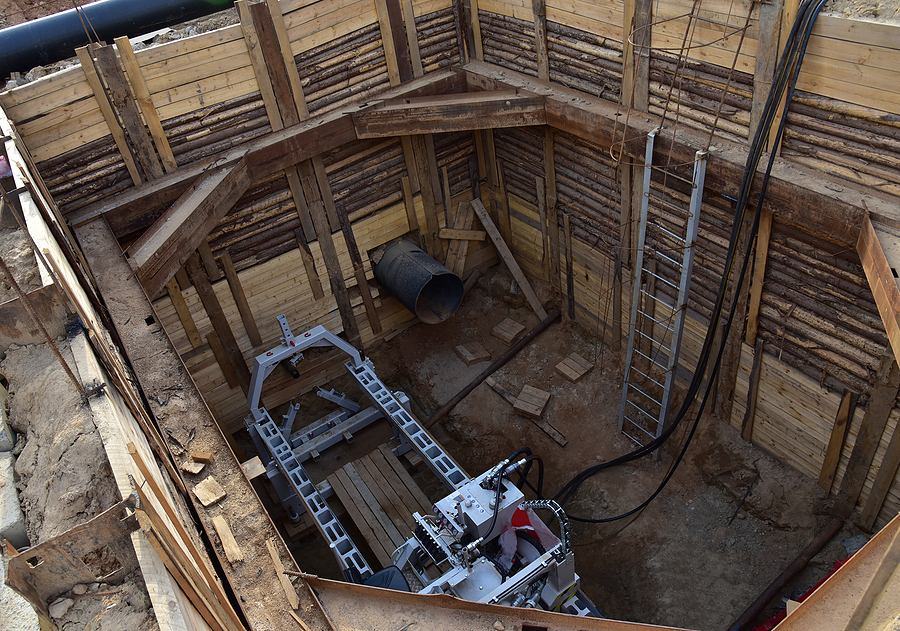 Deep excavation site reinforced with timber shoring, featuring a horizontal boring machine and a section of steel pipe at the base. The setup includes structural supports, a metal ladder, electrical cables, and construction debris, indicating preparation for underground pipe installation or tunneling work.