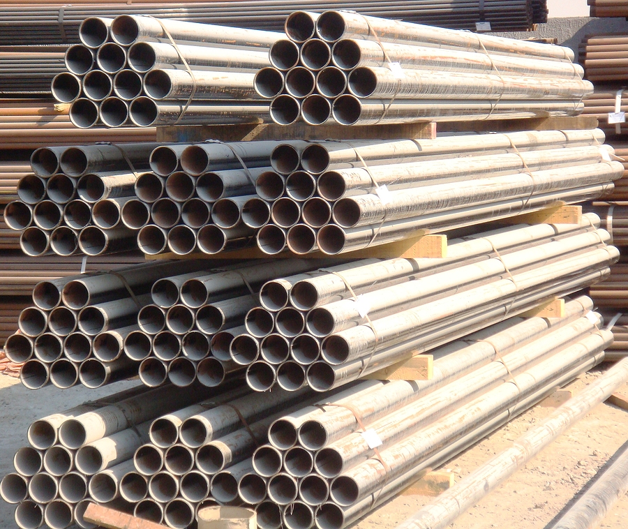 Neatly stacked bundles of new steel pipes organized on wooden supports at an outdoor industrial storage yard. The pipes are arranged in hexagonal formations and secured with metal bands, ready for transport or construction use.